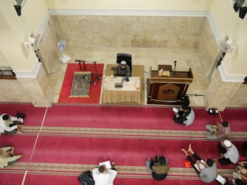 A group of people are seated on red and gold patterned prayer rugs in a large indoor space with cream-colored walls. A man is seated at the front behind a table with microphones, a book, and a cup, indicating he might be delivering a lecture or sermon. A wooden pulpit and a standing fan are nearby.