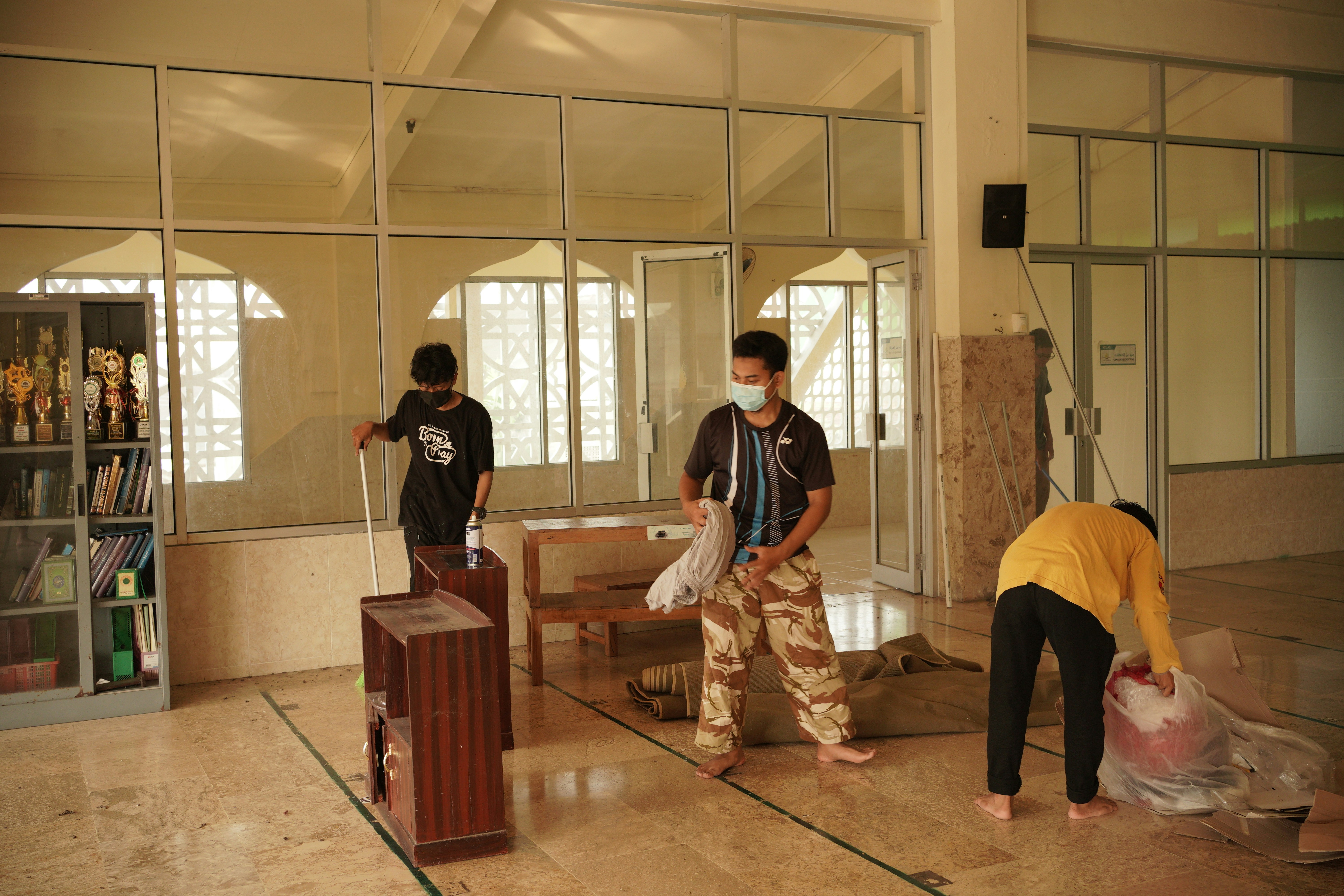 Three individuals are cleaning and arranging items in a room with large glass windows and cabinets filled with trophies and books. One person is sweeping, another is arranging items, and the third is handling a large bag.