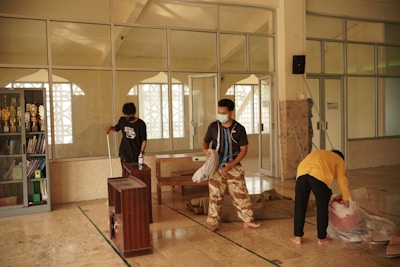 Three individuals are cleaning and arranging items in a room with large glass windows and cabinets filled with trophies and books. One person is sweeping, another is arranging items, and the third is handling a large bag.