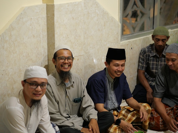 Several men are seated on the floor against a tiled wall, appearing relaxed and smiling. The group is engaged in a social gathering in a warmly lit indoor space. They wear traditional clothing, including caps and sarongs.