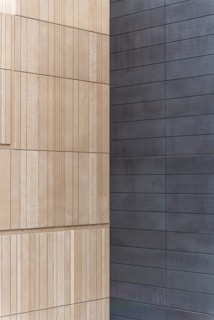 Two walls joined at a corner, with one side featuring light beige, textured rectangular panels arranged in a vertical pattern, and the other side displaying smooth, dark gray horizontal panels.