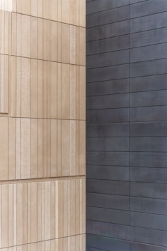 Two walls joined at a corner, with one side featuring light beige, textured rectangular panels arranged in a vertical pattern, and the other side displaying smooth, dark gray horizontal panels.