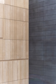 Two walls joined at a corner, with one side featuring light beige, textured rectangular panels arranged in a vertical pattern, and the other side displaying smooth, dark gray horizontal panels.
