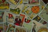 A collection of tarot cards is scattered across a surface. The cards feature various illustrations, including figures, symbols, and numbers, with a vintage and artistic style. The images include the Wheel of Fortune, a knight, a bearded king, and other mystical symbols.