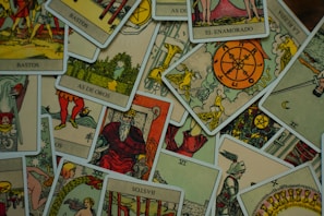 A collection of tarot cards is scattered across a surface. The cards feature various illustrations, including figures, symbols, and numbers, with a vintage and artistic style. The images include the Wheel of Fortune, a knight, a bearded king, and other mystical symbols.