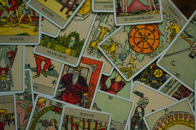 A collection of tarot cards is scattered across a surface. The cards feature various illustrations, including figures, symbols, and numbers, with a vintage and artistic style. The images include the Wheel of Fortune, a knight, a bearded king, and other mystical symbols.