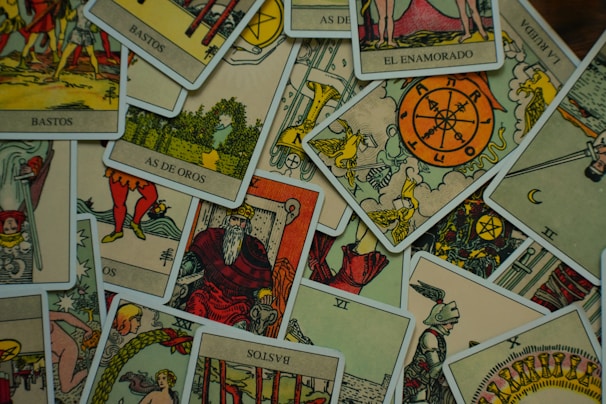 A collection of tarot cards is scattered across a surface. The cards feature various illustrations, including figures, symbols, and numbers, with a vintage and artistic style. The images include the Wheel of Fortune, a knight, a bearded king, and other mystical symbols.