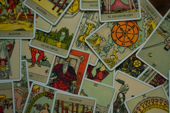 A collection of tarot cards is scattered across a surface. The cards feature various illustrations, including figures, symbols, and numbers, with a vintage and artistic style. The images include the Wheel of Fortune, a knight, a bearded king, and other mystical symbols.