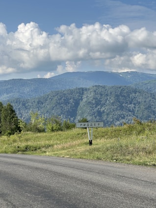 A scenic landscape shows a lush green field adjacent to a paved road with a sign displaying text in Cyrillic. In the background, a series of rolling, tree-covered hills under a partly cloudy blue sky create a serene natural setting.