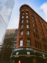 A historic red-brick hotel building stands prominently against a backdrop of modern skyscrapers. The structure features rounded corners and numerous windows, with a green canopy over the entrance that reads 'Palace Hotel'. The sky is bright with scattered clouds.
