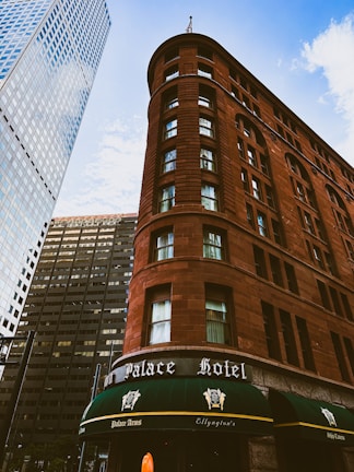A historic red-brick hotel building stands prominently against a backdrop of modern skyscrapers. The structure features rounded corners and numerous windows, with a green canopy over the entrance that reads 'Palace Hotel'. The sky is bright with scattered clouds.