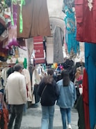 A bustling market corridor filled with various colorful garments hanging on both sides, including dresses, shirts, and traditional wear. The space is crowded with shoppers walking through, and the ceiling of the corridor is adorned with arched stone structures.