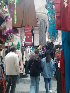A bustling market corridor filled with various colorful garments hanging on both sides, including dresses, shirts, and traditional wear. The space is crowded with shoppers walking through, and the ceiling of the corridor is adorned with arched stone structures.