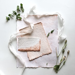 A delicate arrangement featuring textured handmade paper with frayed edges, set alongside sprigs of fresh herbs and twigs. A sheer white ribbon meanders through the composition, adding a soft, elegant touch.