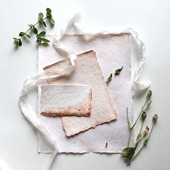 A delicate arrangement featuring textured handmade paper with frayed edges, set alongside sprigs of fresh herbs and twigs. A sheer white ribbon meanders through the composition, adding a soft, elegant touch.