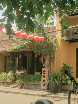 A quaint street scene features a building with a yellow facade and a tiled roof. Red lanterns are strung across the front of the building, adding a vibrant contrast. Greenery and potted plants line the sidewalk, while dappled sunlight filters through overhead tree branches. A wooden sign displays 'CHOU Kitchen & Bar.'