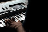 A person is playing a keyboard with visible notes and controls on the instrument panel. The hand is positioned over the keys, and the lighting is dim, suggesting a focused and intimate setting. The keyboard has a digital display and various buttons and sliders for musical adjustments.