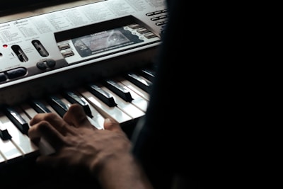 A person is playing a keyboard with visible notes and controls on the instrument panel. The hand is positioned over the keys, and the lighting is dim, suggesting a focused and intimate setting. The keyboard has a digital display and various buttons and sliders for musical adjustments.