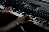 A pair of hands skillfully playing an electronic keyboard. The keys are black and white, and there are various buttons and a display screen on the keyboard for controlling sounds and settings. The lighting is dim, creating a focused and intimate mood.