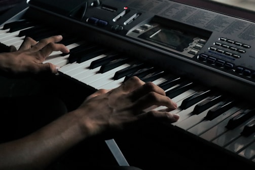 A pair of hands skillfully playing an electronic keyboard. The keys are black and white, and there are various buttons and a display screen on the keyboard for controlling sounds and settings. The lighting is dim, creating a focused and intimate mood.