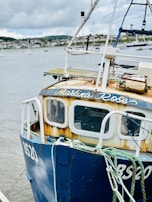 A weathered blue fishing boat with the name 'Martina Rose' on its cabin stands docked at a harbor. The boat exhibits signs of rust and aging. Several ropes are secured to its side. In the background, multiple sailboats are anchored on a calm body of water, with a distant shoreline visible under a cloudy sky.