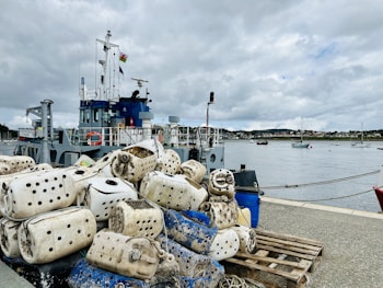 A harbor scene with a vessel anchored at the dock, featuring a stack of large, worn, cylindrical floats and buoys piled on wooden pallets. The horizon shows a calm body of water with more boats in the distance under an overcast sky.
