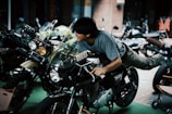 A person is playfully lying on a motorcycle, holding onto the handlebars, while other motorcycles are parked nearby. The setting appears to be an indoor or covered space, with lighting creating a somewhat dramatic effect.