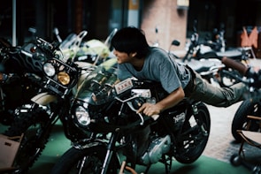 A person is playfully lying on a motorcycle, holding onto the handlebars, while other motorcycles are parked nearby. The setting appears to be an indoor or covered space, with lighting creating a somewhat dramatic effect.