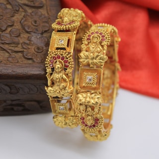 Two intricately designed gold bangles, adorned with detailed carvings of deities and floral motifs. The bangles feature small embedded red stones and square diamond-like embellishments. A wooden box with floral patterns is partially visible in the background, set against a soft red fabric.