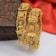 Two intricately designed gold bangles, adorned with detailed carvings of deities and floral motifs. The bangles feature small embedded red stones and square diamond-like embellishments. A wooden box with floral patterns is partially visible in the background, set against a soft red fabric.