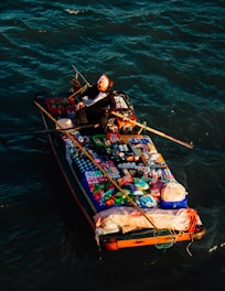 A small boat is floating on water, loaded with a variety of goods including food items, toiletries, and other consumables. There is a person sitting on the boat, rowing with oars. The person is wearing a black outfit and has a white cloth around their head.