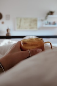 A warm-toned image of a hand holding a cream-colored mug against a backdrop of a cozy, sunlit living room.