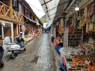 A narrow street is lined with small shops displaying a variety of colorful textiles, souvenirs, and decorative items. Two scooters are parked on the cobblestone path, which is wet, suggesting recent rain. The shops have wooden facades and are covered with tarps, with various goods hanging outside, including necklaces and small statues. A few people are visible in the background, walking through the market area.