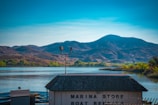 A serene lake reflecting the blue sky and surrounded by mountains in the distance. The foreground features a building with a shingled roof and a sign indicating a marina store and boat rentals. Vegetation is visible along the water's edge, adding a splash of green to the scene.
