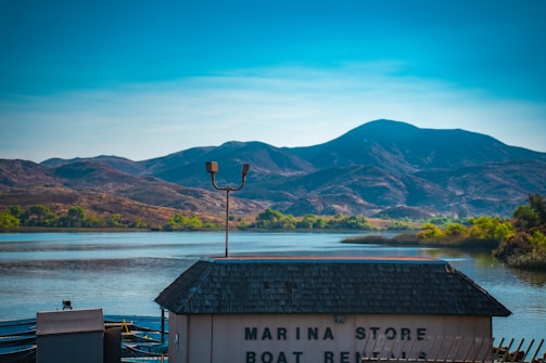 A serene lake reflecting the blue sky and surrounded by mountains in the distance. The foreground features a building with a shingled roof and a sign indicating a marina store and boat rentals. Vegetation is visible along the water's edge, adding a splash of green to the scene.
