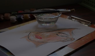 A watercolor painting depicts a serene face on a piece of paper resting on a table. Nearby, a glass cup filled with clear water holds two paintbrushes, and small paint containers are scattered around.