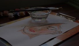 A watercolor painting depicts a serene face on a piece of paper resting on a table. Nearby, a glass cup filled with clear water holds two paintbrushes, and small paint containers are scattered around.