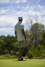 A person wearing a patterned shirt, shorts, a cap, and golf shoes is holding a golf club and standing on a golf course. The background features lush greenery and cloudy skies, suggesting a peaceful outdoor setting.