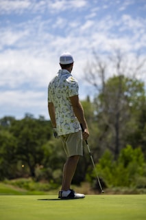 A person wearing a patterned shirt, shorts, a cap, and golf shoes is holding a golf club and standing on a golf course. The background features lush greenery and cloudy skies, suggesting a peaceful outdoor setting.