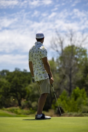 A person wearing a patterned shirt, shorts, a cap, and golf shoes is holding a golf club and standing on a golf course. The background features lush greenery and cloudy skies, suggesting a peaceful outdoor setting.