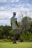 A person wearing a patterned shirt, shorts, a cap, and golf shoes is holding a golf club and standing on a golf course. The background features lush greenery and cloudy skies, suggesting a peaceful outdoor setting.