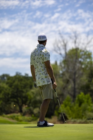 A person wearing a patterned shirt, shorts, a cap, and golf shoes is holding a golf club and standing on a golf course. The background features lush greenery and cloudy skies, suggesting a peaceful outdoor setting.