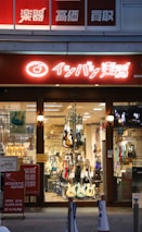 A storefront displaying a variety of guitars, both electric and acoustic, in a well-lit shop. The store has a large window through which several guitars are visible hanging or standing. A brightly lit sign with Japanese characters is above the entrance, and posters with additional information are displayed near the entrance.