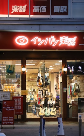 A storefront displaying a variety of guitars, both electric and acoustic, in a well-lit shop. The store has a large window through which several guitars are visible hanging or standing. A brightly lit sign with Japanese characters is above the entrance, and posters with additional information are displayed near the entrance.