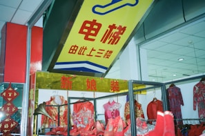 A clothing store display featuring traditional Chinese attire, including dresses and shoes in vibrant red and gold colors. The shop has a prominent sign in Chinese characters above the display, and the interior ceiling is visible with recessed lighting.