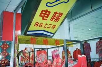 A clothing store display featuring traditional Chinese attire, including dresses and shoes in vibrant red and gold colors. The shop has a prominent sign in Chinese characters above the display, and the interior ceiling is visible with recessed lighting.