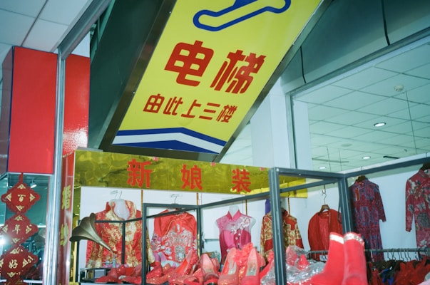 A clothing store display featuring traditional Chinese attire, including dresses and shoes in vibrant red and gold colors. The shop has a prominent sign in Chinese characters above the display, and the interior ceiling is visible with recessed lighting.