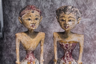 Two aged, weathered wooden sculptures are seen, each with distinct facial features and detailed carvings. They have a worn appearance, with visible paint erosion, particularly around the face and body. Both sculptures are dressed in what appears to be traditional attire, characterized by faded red and beige colors.