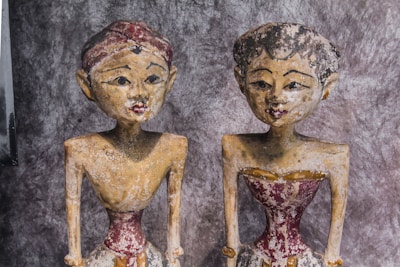 Two aged, weathered wooden sculptures are seen, each with distinct facial features and detailed carvings. They have a worn appearance, with visible paint erosion, particularly around the face and body. Both sculptures are dressed in what appears to be traditional attire, characterized by faded red and beige colors.