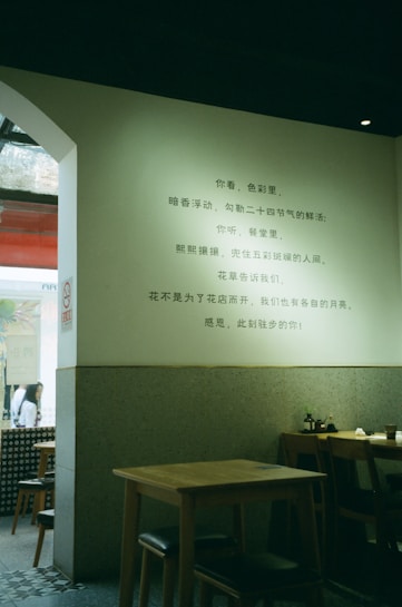 A cozy interior of a restaurant with minimalist wooden tables and chairs is shown. A poem or text in Chinese is written on the light-colored wall. The setting appears serene and quiet, with soft lighting enhancing the tranquil atmosphere.